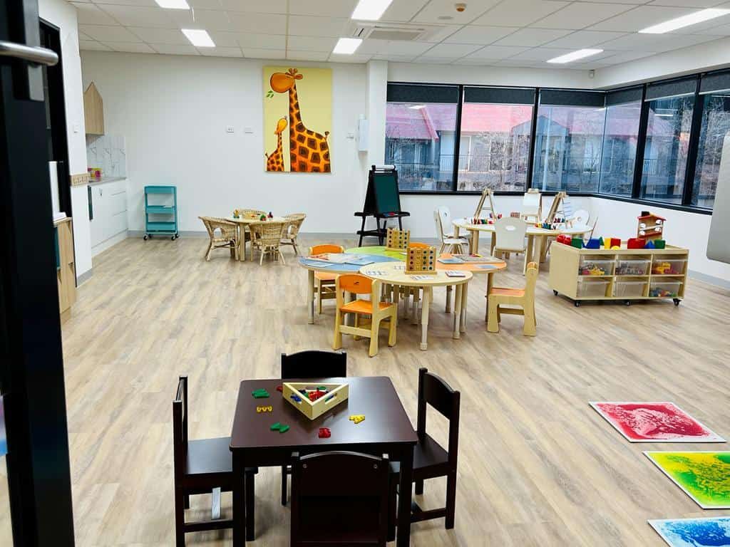 A bright toddler room with a giraffe artwork, small tables and chairs and sensory floor tiles