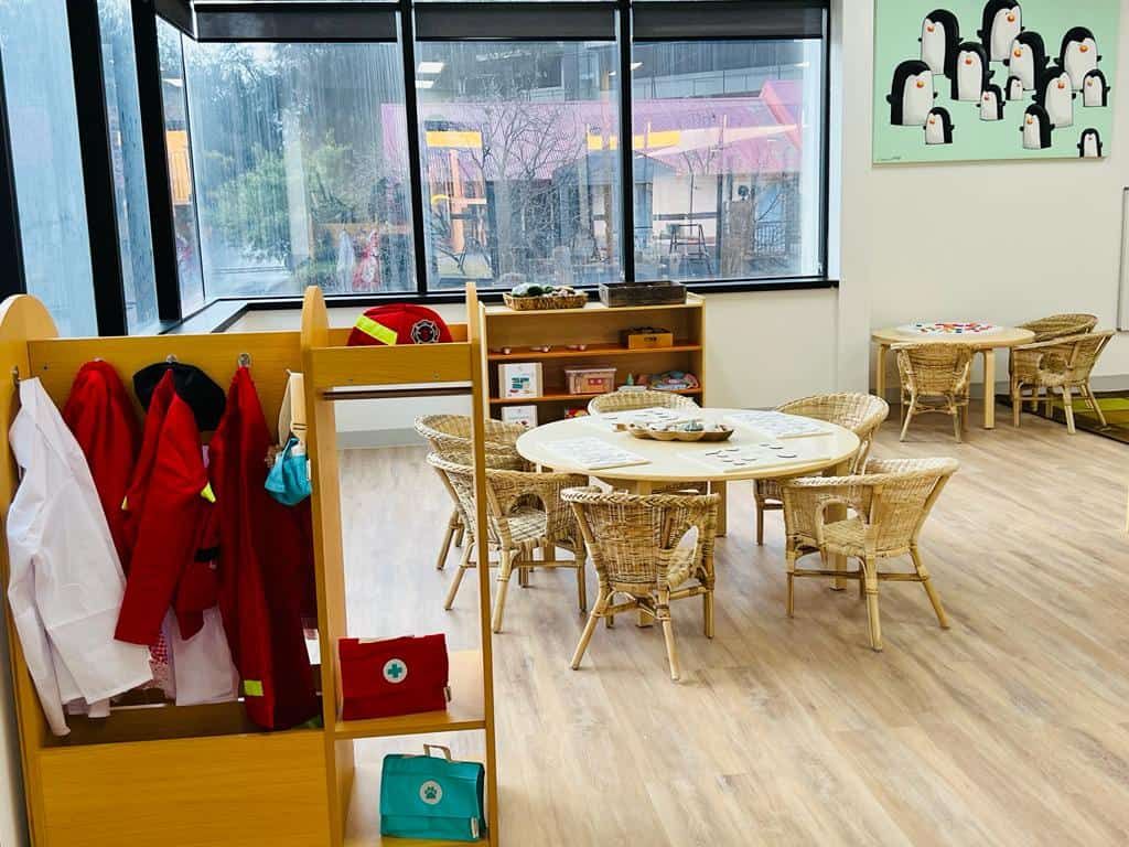 A preschool room with a penguin artwork, dress-up clothes and round tables with cane chairs
